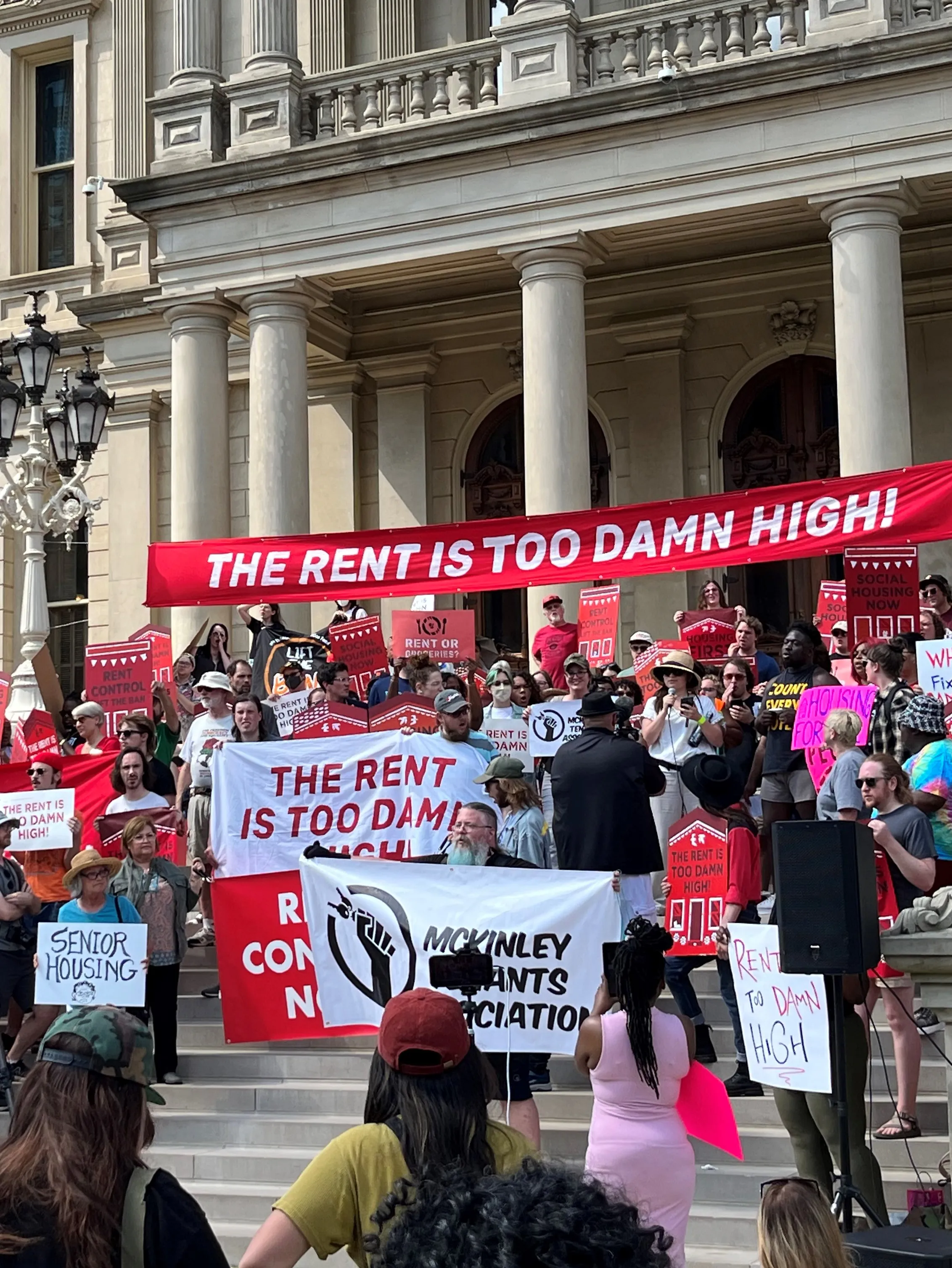 a giant rally for fairer rents fills a public plaza. many hold signs which read 'THE RENT IS TOO DAMN HIGH'